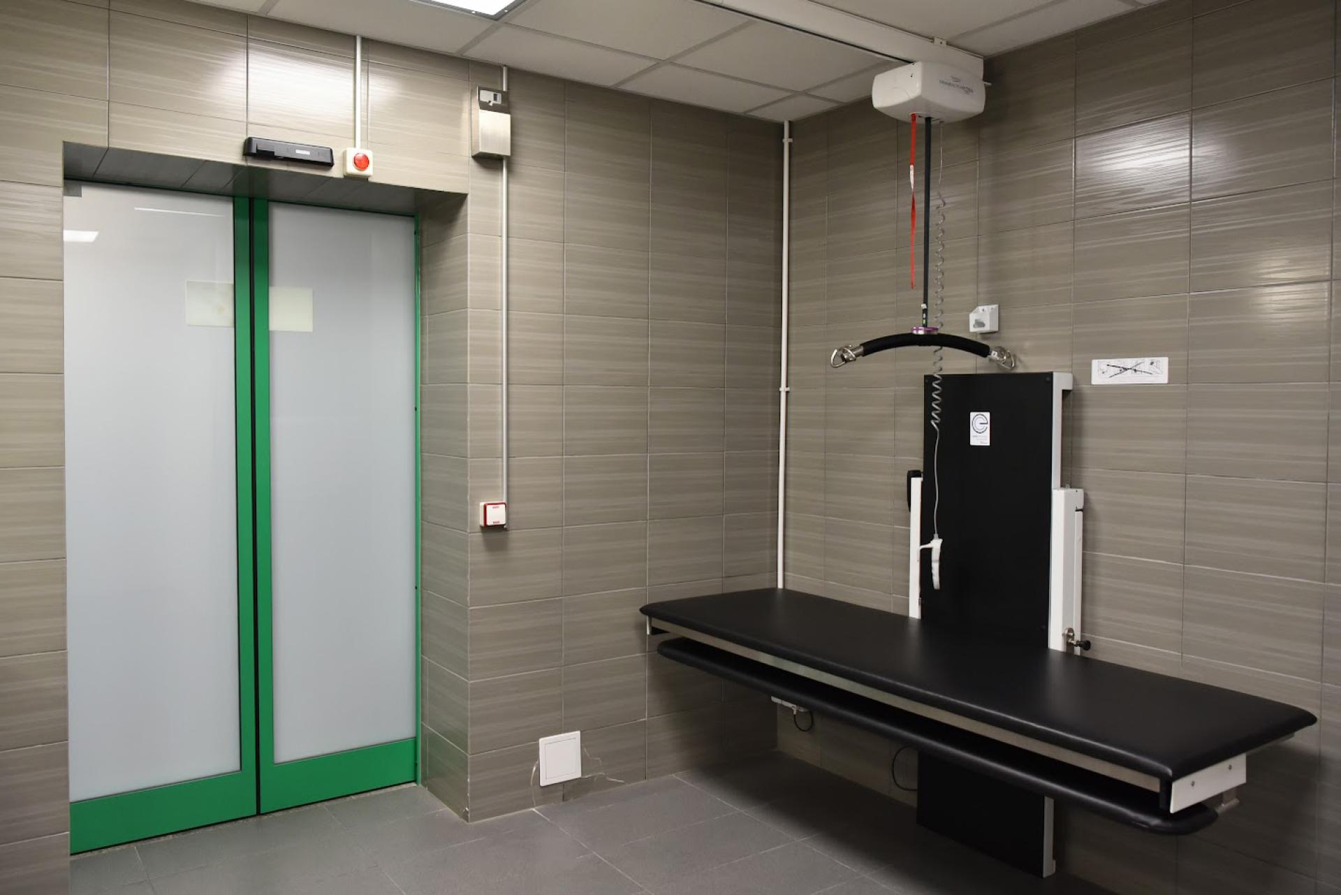 Zdjęcie przedstawia fragment pomieszczenia, w którym zainstalowano regulowaną leżankę oraz elektryczny podnośnik.