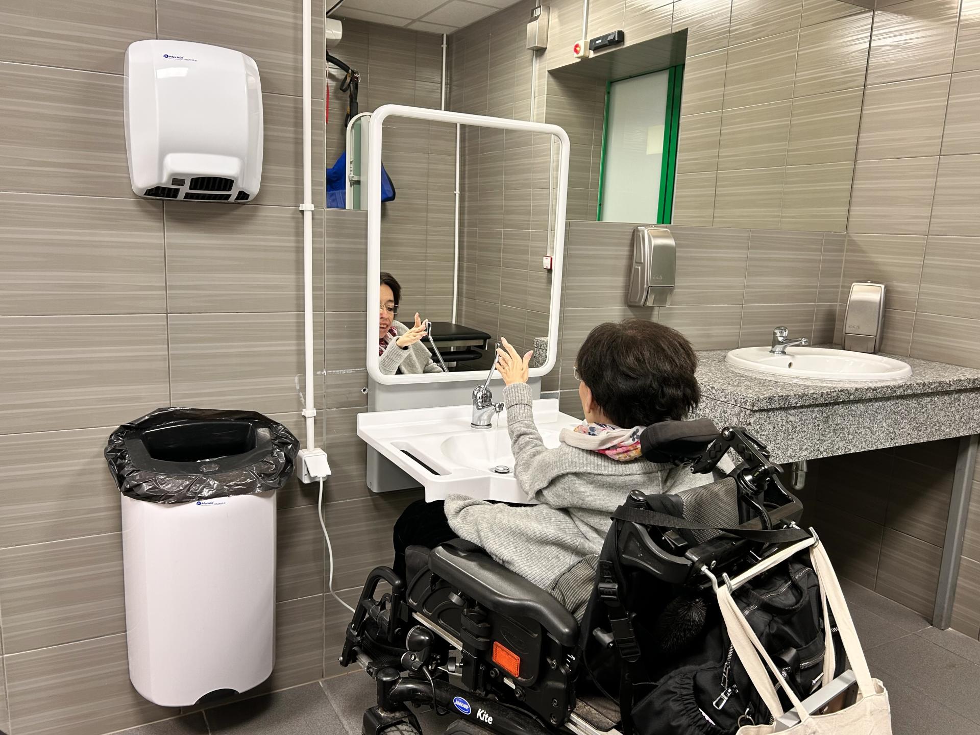 Kobieta na wózku przed jedną z umywalek - ruchomą, regulowaną wraz z lustrem. Z prawej strony nieruchoma umywalka w kamiennym blacie, z lewej - duży kosz na odpadka nad nim suszarka do rąk.k