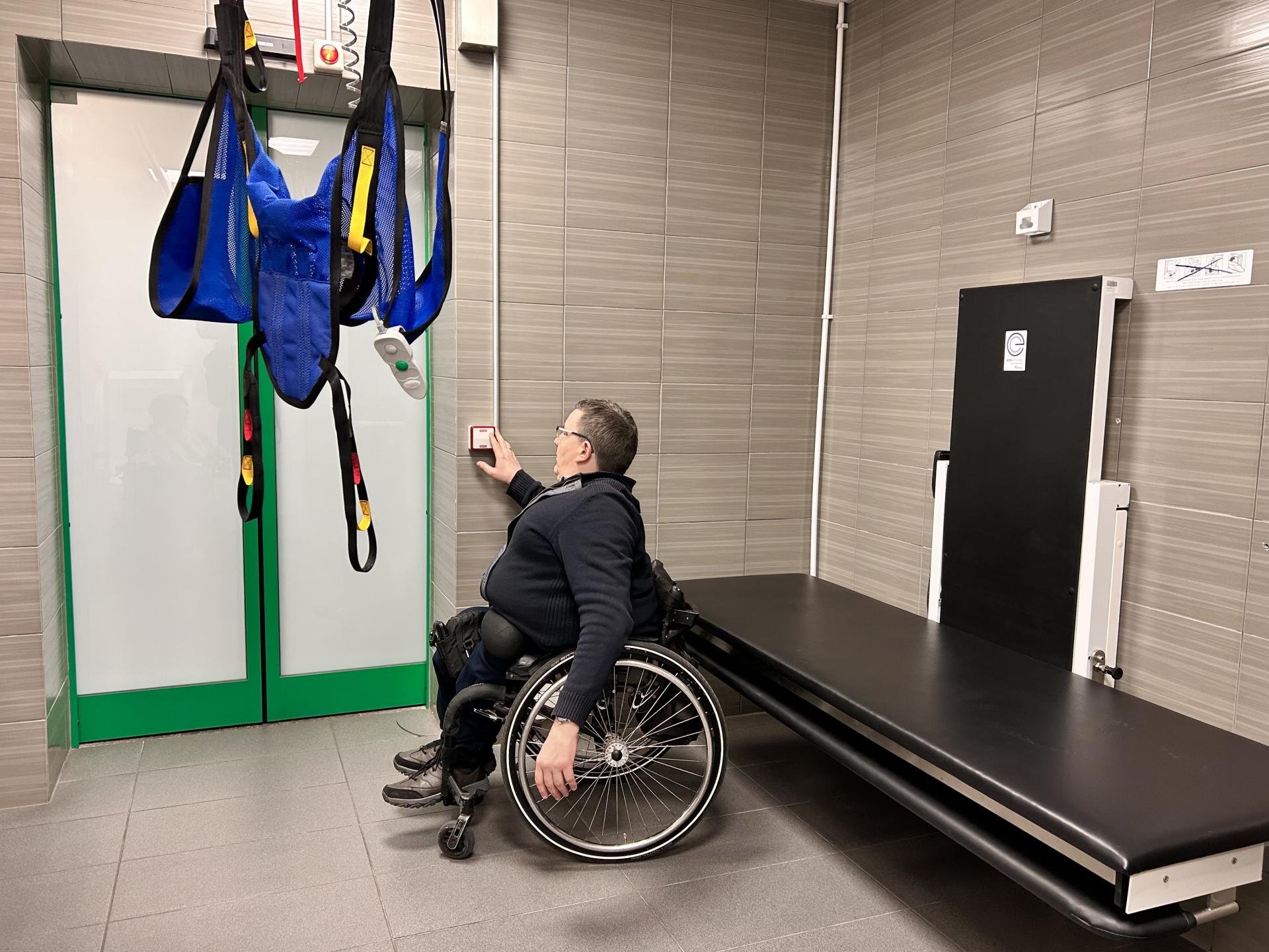 Mężczyzna na wózku opuszcza nosidełko podnośnika. Za nim czarna leżanka, regulowana elektrycznie.