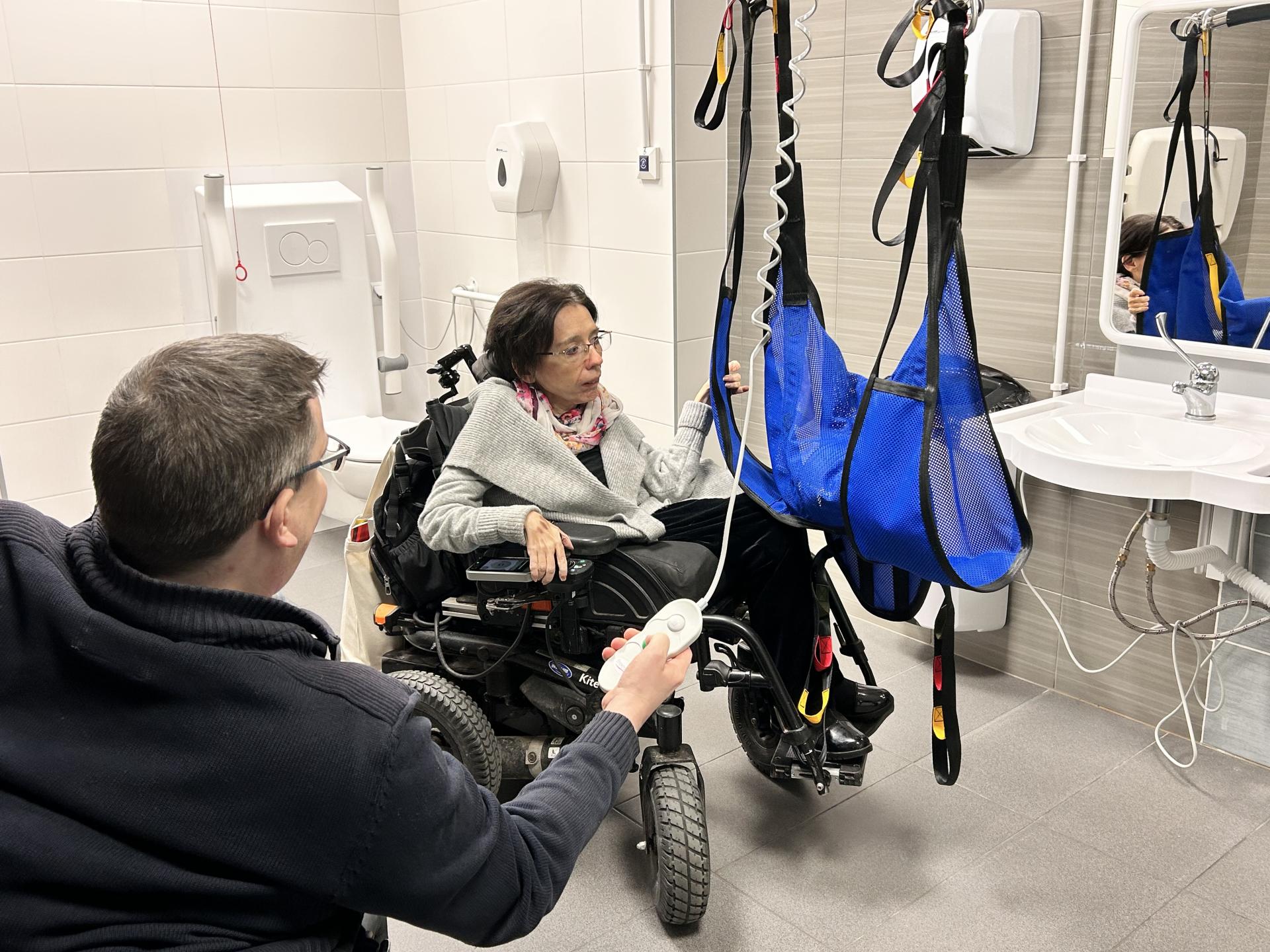 Dwie osoby na wózkach, kobieta i mężczyzna, testują w komfortce podnośnik i nosidełko. Za nimi sedes z widoczną linką alarmowa, z prawej strony zdjęcia ruchoma umywalka z lustrem.