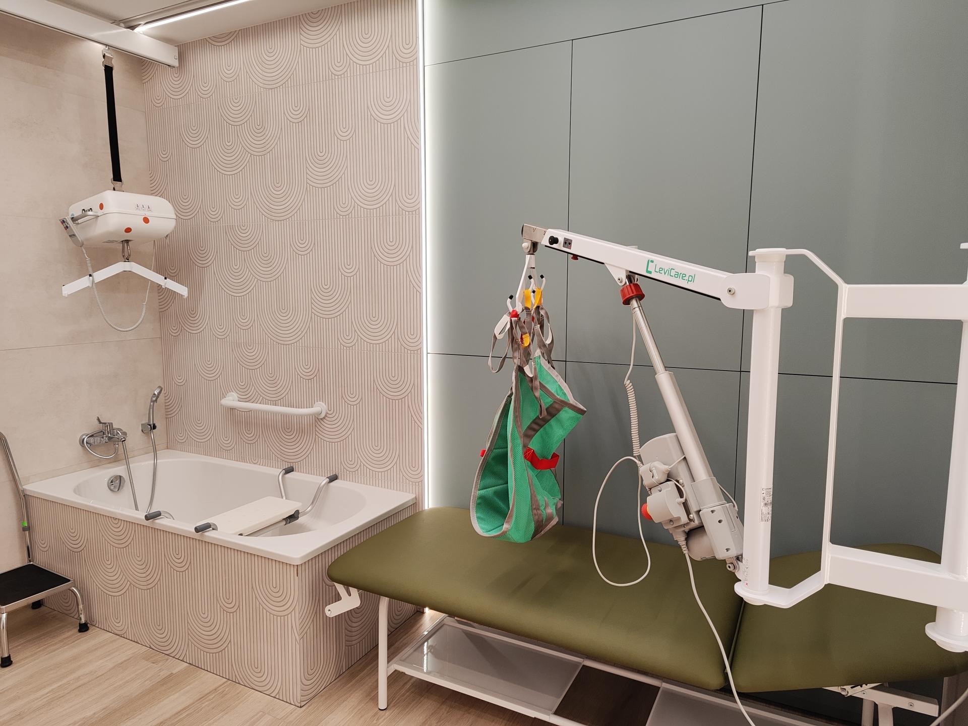We wnętrzu komfortki znajdują się leżanka z podnośnikiem bocznym, a obok wanna z podnośnikiem sufitowym