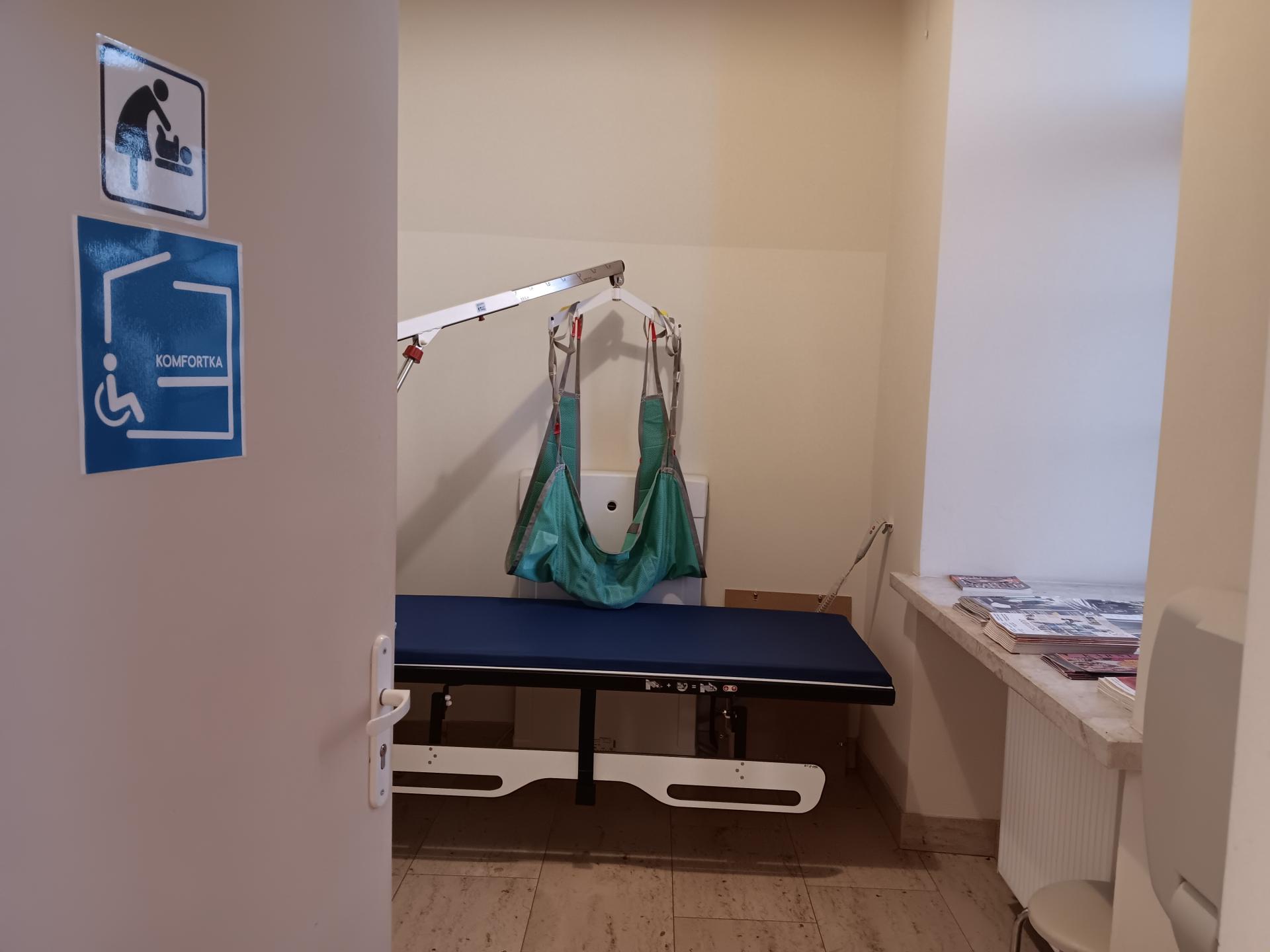 Wnętrze komfortki zregulowaną leżanką i podnośnikiem przyściennym. Widok przez uchylone drzwi z piktogramem komfortki i pokoju dla rodzica z dzieckiem. To komfortka na Wawelu.
