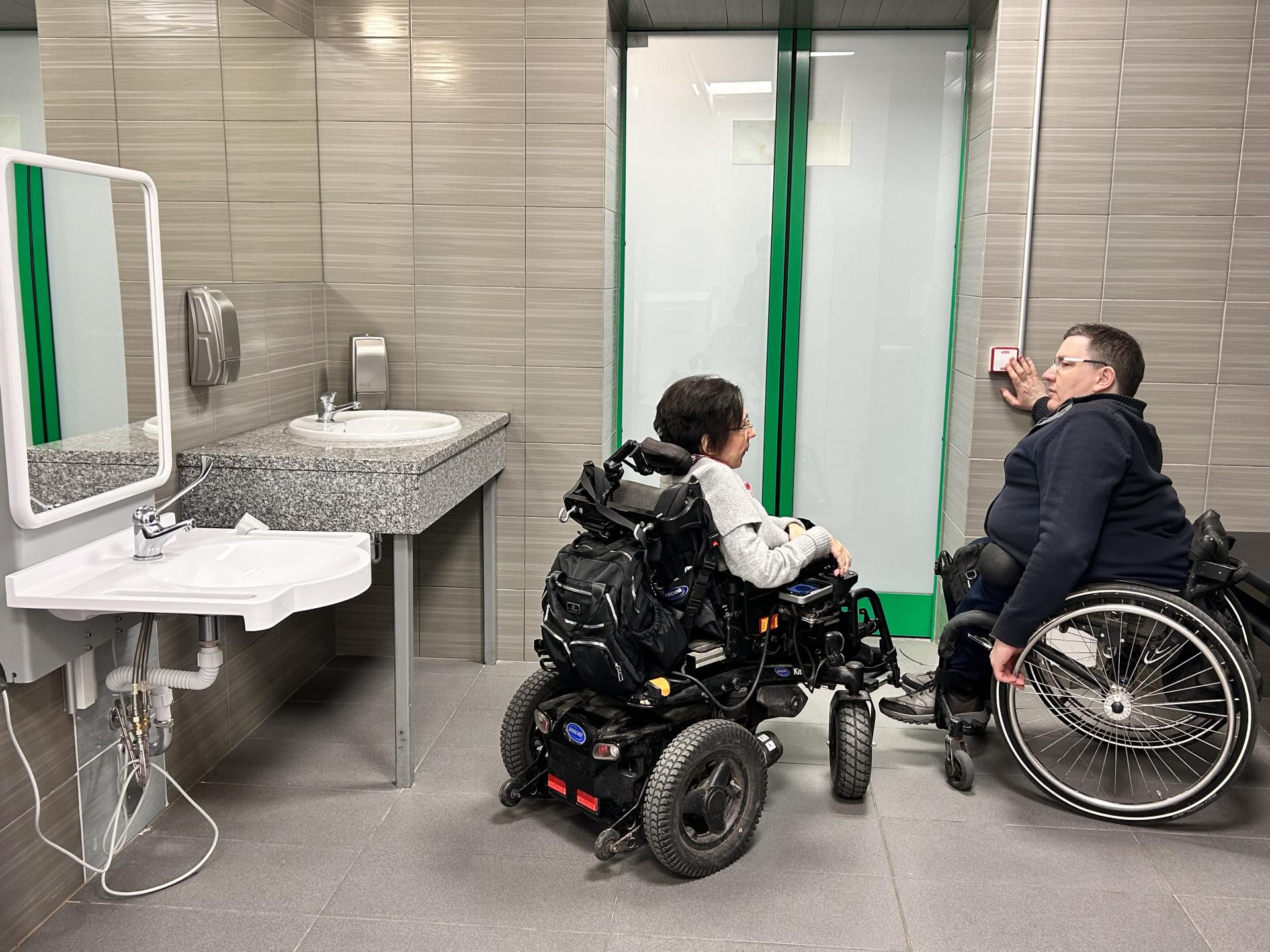 Dwie soby na wózkach we wnętrzu komfortki, Widać drzwi, regulowaną i nieregulowaną umywalkę i lustro