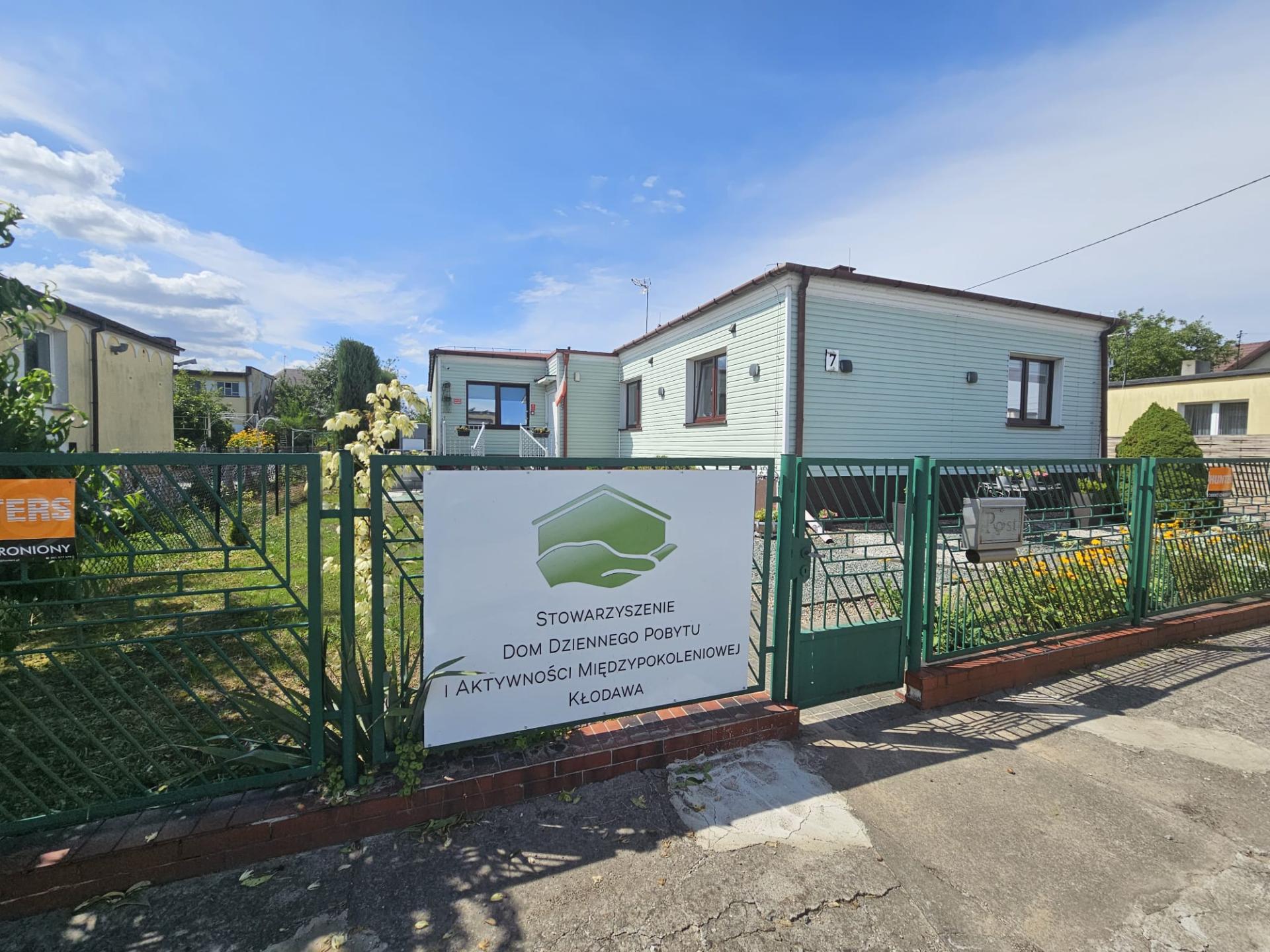 Parterowy, jasny budynek ogrodzony zielonym metalowym płotem z planszą: Stowarzyszenie Dom Dziennego Pobytu i Aktywności Międzypokoleniowej w Kłodawie
